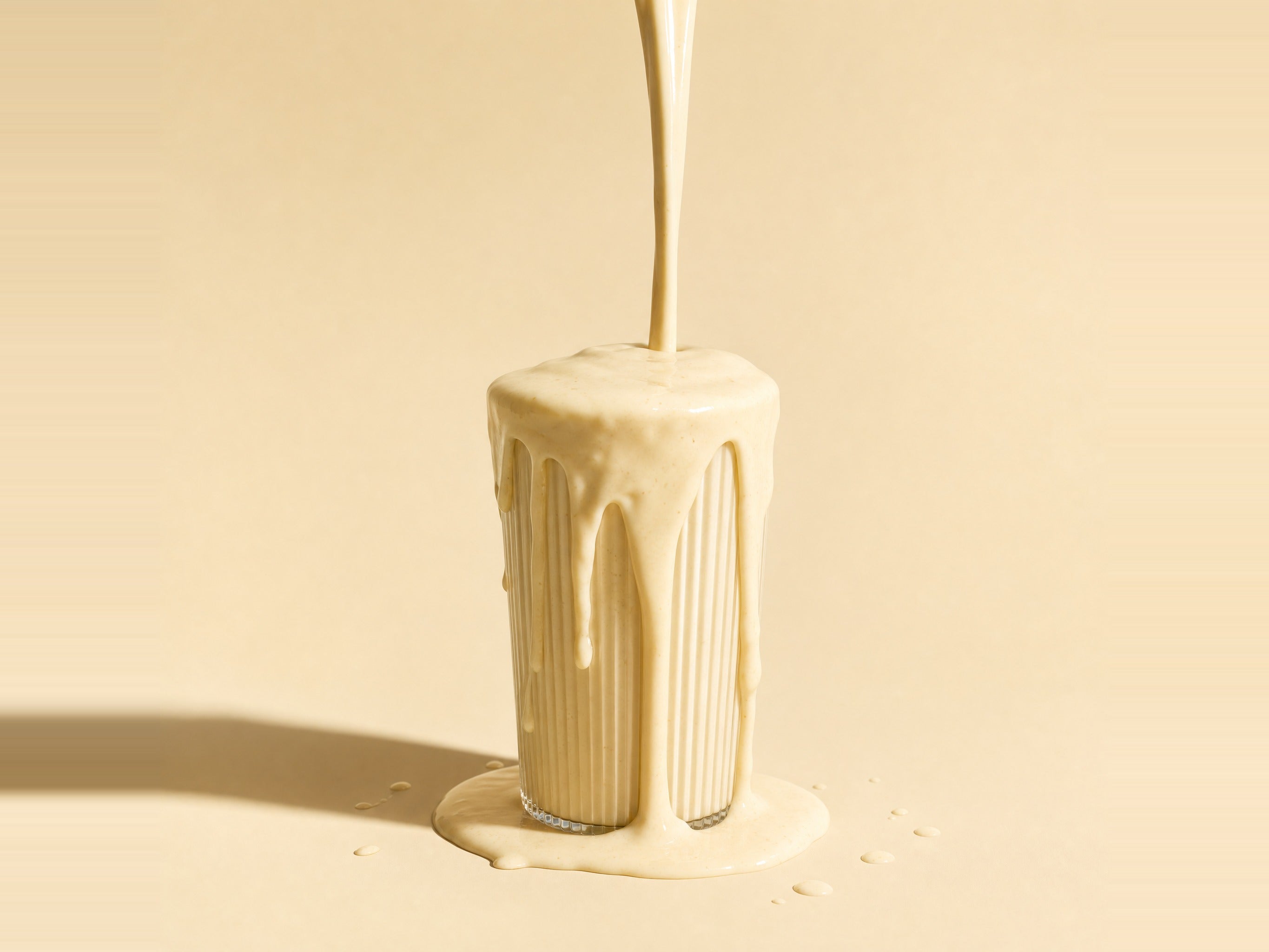 Creamy Heey! Protein & Biotics vanilla shake overflowing a ribbed glass on a warm cream backdrop